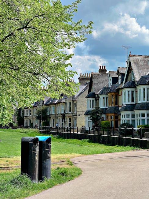 The image depicts a quiet residential street with a row of traditional terraced houses featuring bay windows, slate roofs, and chimneys, situated behind a low black metal fence. In the foreground, there are two black upright bins positioned on the pavement near a grassy area, which appears to be part of a public park or communal space. A large leafy tree with fresh green foliage extends its branches over the scene, providing shade and adding a natural element to the setting. The background showcases a blue sky with scattered white clouds, suggesting a clear day. The scene conveys a tidy and well-maintained urban environment, where occasional waste collection or private rubbish disposal by local residents may take place. Overall, the image highlights a typical UK suburban street scene with an emphasis on curbside waste management suited for households serviced by independent rubbish removal services like Waste Disposal Shepherds Bush.