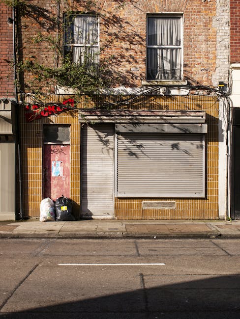 The image depicts the front façade of a small commercial storefront in an urban setting, with a closed grey metal roller shutter covering the shop entrance. To the left of the shutter, there is a narrow metal door, partially painted red with some graffiti, which appears to provide access or emergency exit. Adjacent to the entrance, two black household waste bags and a white plastic bag are placed on the pavement, likely containing rubbish awaiting collection. The building's exterior wall is clad in brownish-yellow tiles, with some signs of wear and weathering. Above, on the upper level, there are two large rectangular windows with white frames and partially drawn curtains, set into a brick façade that shows visible signs of age, such as uneven brickwork and minor graffiti. A vine with sparse green leaves extends across the upper section of the brick wall, casting shadows on the windows. Overhead, a series of black cables runs horizontally, attached to the building, and a small satellite dish is mounted near the center of the upper wall. The scene is illuminated by natural daylight, with shadows suggesting a clear sky, and the curb and street appear clean apart from the visible rubbish bags, consistent with private waste disposal or alternative rubbish collection services in an urban environment.