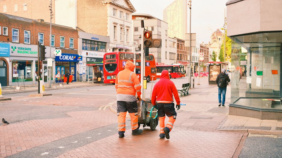 Two workers dressed in high-visibility orange and red safety clothing are pushing a wheeled trash bin along a paved city sidewalk. The worker in orange, facing away, holds the handle of the bin, while the worker in red guides it from behind. Behind them, there are various urban storefronts including a pharmacy, a building society office, and a shop with signage. In the background, red double-decker buses are visible on a busy street, with some pedestrians walking along the sidewalk and a bus stop with an advertisement display on the right. The environment is well-lit with natural daylight, and surrounding buildings show a mix of modern and traditional architecture, contributing to an urban rubbish removal scene typical of city street sanitation operations by Waste Disposal Shepherds Bush or similar private waste management services.